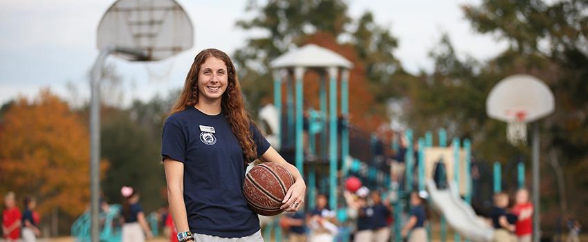Female holding basketball in front of outdoor basketball court and playground.