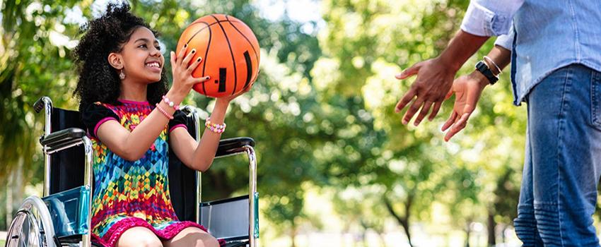 Young girl in wheelchair playing basketball with an adult.